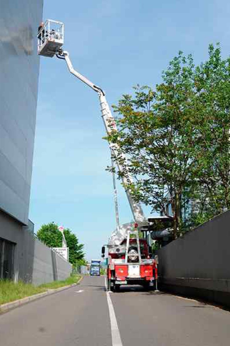 Rote ausgefahrene Arbeitsbühne zwischen zwei gläsernen Gebäuden, rechts ein Baum.