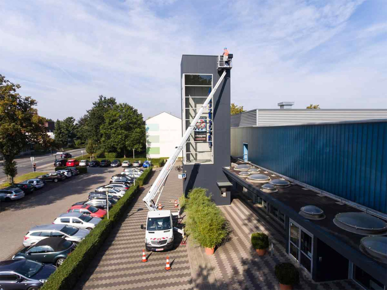 Links Bäume, Parkplatz mit Autos; rechts Gebäude und ausgefahrene Arbeitsbühne.
